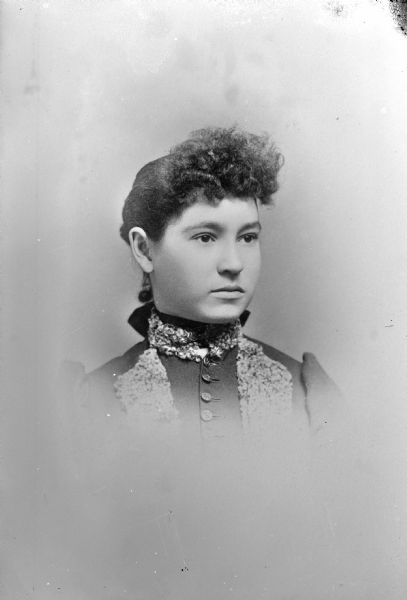 Vignetted head and shoulders studio portrait of an unidentified European American woman posed sitting. She is wearing a dark-colored dress with a button-down bodice, with light-colored thick trim at neck and at bodice. She is also wearing a collar pin.