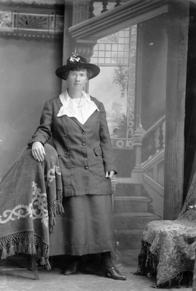 Full-length studio portrait in front of a painted backdrop of an unidentified European American woman posed standing with her right hand resting on the back of a chair draped with a tasseled cloth. She is wearing a dark-colored skirt, three-quarter length loose-fitting jacket, a hat with ribbon and trim, a light-colored blouse with large collar, and a collar pin.