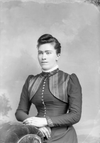 Waist-up studio portrait of an unidentified European American woman posed standing with her hands resting on the back of an upholstered chair. She is wearing a dark-colored dress with a high collar, collar pin, button-down bodice, and striped bolero-style detail at collar and shoulders.