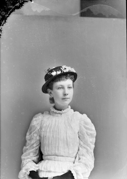 Waist-up studio portrait of an unidentified young European American girl posed sitting. She is wearing a light-colored pleated dress, with a sheer fabric overlay decorated with white dots. She is also wearing dark-colored fingerless gloves, dark-colored straw hat with flowers, earrings, a choker necklace, and a small collar pin.
