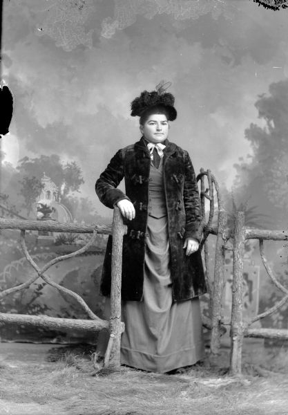Full-length studio portrait of an unidentified European American woman posed standing in front of a painted backdrop in the opening of a gate on a fence. She is wearing an open, three-quarter length heavy coat with toggle buttons, basque bodice and gathered overskirt, and a hat decorated with dark ostrich feathers.