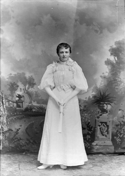 Full-length studio portrait in front of a painted backdrop of an unidentified young European American woman posed standing. She is holding a diploma with both hands in front of her, and is wearing over the elbow length gloves, a light-colored full-length dress, and white low-heeled shoes. There is ribbon detail on the shoulders and belt of the dress, which has puffy sleeves. This may be for a high school graduation.