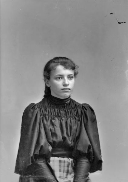 Waist-up studio portrait of an unidentified young European American woman posed sitting. She is wearing a plaid skirt and a dark-colored, smocked blouse with puffy sleeves.