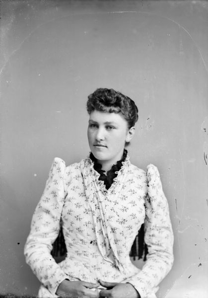 Waist-up studio portrait of an unidentified European American woman posed sitting in a chair with a tasseled back. She is wearing a light-colored floral print dress with a dark lace collar.