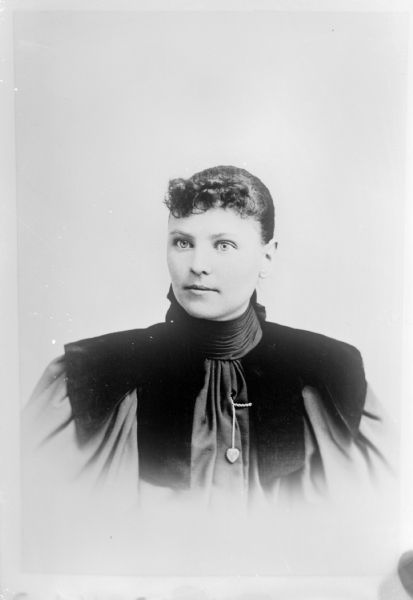 Copy photograph of a quarter-length studio portrait of an unidentified European American woman posed sitting. She is wearing a dark-colored dress with puffy sleeves, dark trim at shoulders and along sides of bodice, and a heart-shaped buttonhole pendant.