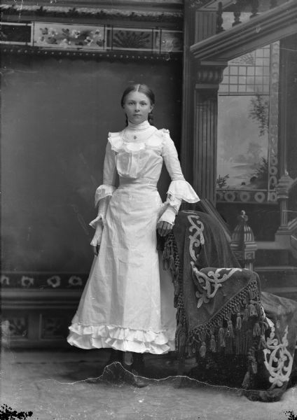 Full-length studio portrait in front of a painted backdrop of an unidentified young European American woman posed standing next to a chair covered with an embroidered and tasseled cloth. She is wearing a crisp, white dress with a high collar, lace details at bodice, elbows and cuffs, and ruffles at the bottom of the skirt, which ends at the top of her high-button shoes. She is wearing a necklace or pin, and white fingerless gloves. Her center-parted hair is pulled back in two braids.