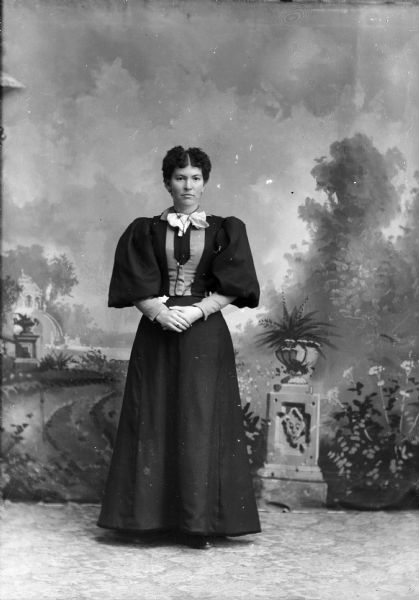 Full-length studio portrait in front of a painted backdrop of an unidentified European American woman posed standing. She is wearing a dark-colored dress with light-colored trim at bodice and at wrists. The sleeves are puffy and full from the shoulder to the elbow. She also wears a ribbon bow neckerchief, and a cord around her neck that is probably for a pocket watch tucked at her waist.