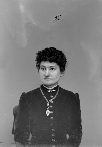 Waist-up studio portrait of a European American woman posed sitting in a chair with a tasseled back. She is wearing a dark-colored, perhaps velvet, button-down bodice, a collar pin, and a heavy chain necklace with locket.
