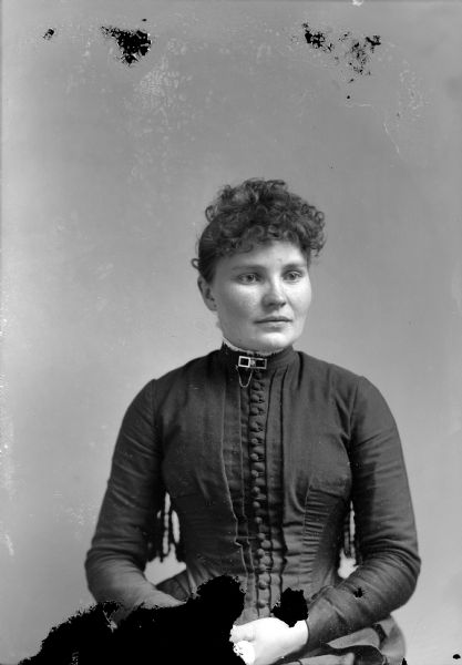 Waist-up studio portrait of an unidentified European American woman posed sitting in a chair with a tasseled back. She is wearing a dark-colored tight-fitting button-down bodice and a collar pin with a chain. 	