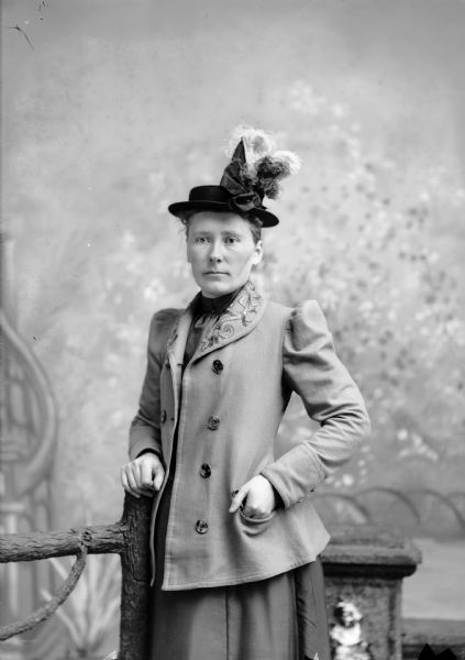 Three-quarter length studio portrait in front of a painted backdrop of an unidentified European American woman posed standing. She has her right hand on a prop wooden fence and her left hand in her pocket. She is wearing a light-colored jacket with embroidery on the lapels, a dress and a hat decorated with a dark bow and ostrich feathers.