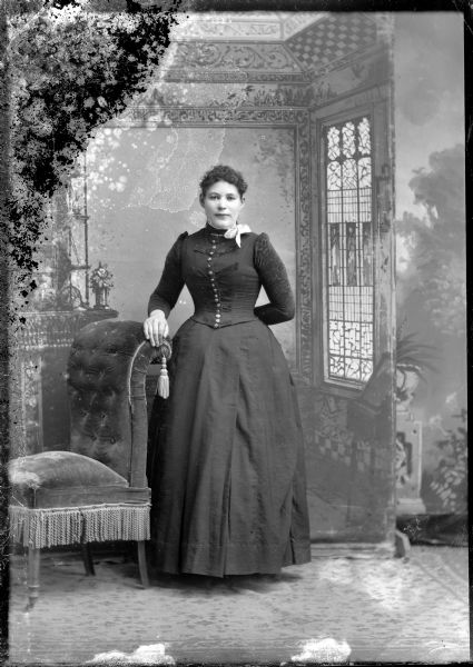 Full-length studio portrait in front of a painted backdrop of an unidentified European American woman posed standing. She has her right hand resting on the back of an upholstered chair, and her left arm is behind her back. She is wearing a dark-colored dress with a button-down bodice, a collar pin, and a ribbon neckerchief.