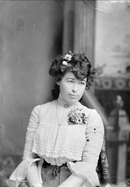 Waist-up studio portrait in front of a painted backdrop of an unidentified European American woman posed sitting. She is wearing a light-colored lace blouse, collar pin, chain necklace, corsage, dark-colored skirt, and a ribbon and flowers in her hair.