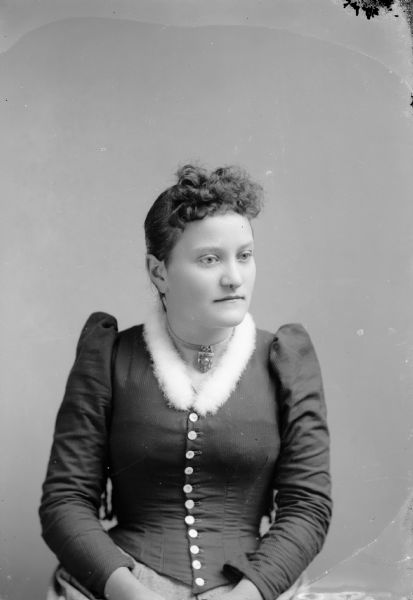 Waist-up studio portrait of an unidentified European American woman posed sitting in a chair with a tasseled back. She is wearing a dark-colored tight-fitting button-down bodice that comes to a point just below her waist. At the collar is a fluffy white fur lining, and she is wearing a flat metal choker with a large pendant.