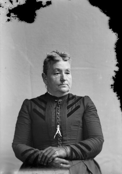 Waist-up studio portrait of an unidentified European American woman posed sitting. She is wearing a dark-colored dress with a pleated button down bodice and darker colored trim, gloves, a buttonhole ribbon and a straight collar pin.