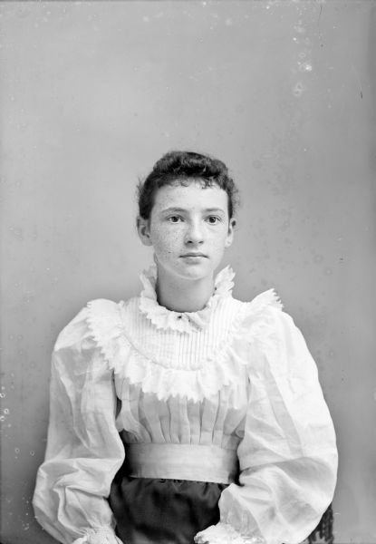 Waist-up studio portrait of a young European American woman posed sitting in a chair with a tasseled back. She wearing a dark-colored skirt, and a white blouse with a pleated and ruffled bodice, and a small collar pin.