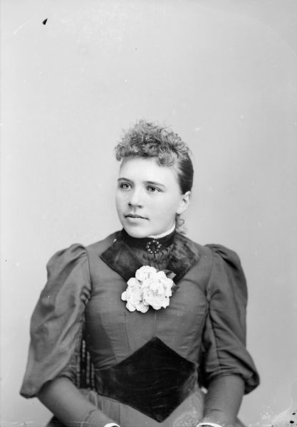 Waist-up studio portrait of a European American woman posed sitting in a chair with a tasseled back. She is wearing a dark-colored dress with dark velvet trim, a round collar pin, and a corsage of flowers.