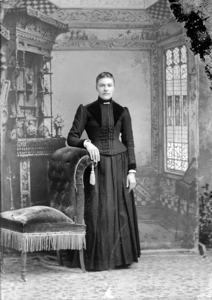 Full-length studio portrait in front of a painted backdrop of an unidentified European American woman posed standing, and resting her right arm on the back of an upholstered chair. She is wearing a dark-colored dress with a pleated bodice and velvet trim, a ring, and a collar pin.