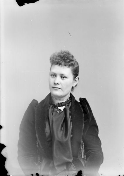 Waist-up studio portrait of an unidentified European American woman posed sitting in a chair. She is wearing a dark-colored blouse, collar pin with chain, and choker necklace.