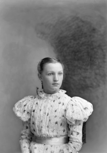 Waist-up studio portrait of an unidentified young European American woman posed sitting in a chair with a tasseled back. She is wearing a light-colored floral print dress with a belt at the waist and puffy sleeves.