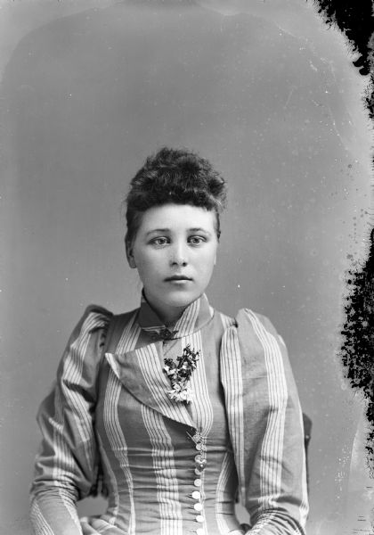 Waist-up studio portrait of an unidentified European American woman posed sitting. She is wearing a light-colored striped dress with a button-down bodice, a diamond collar pin, buttonhole chain with a heart-shaped charm, and a corsage.