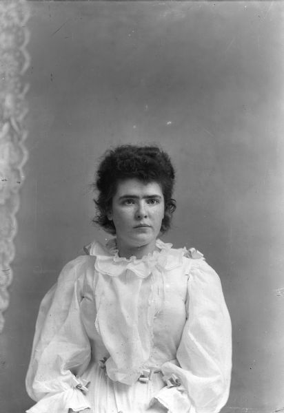 Waist-up studio portrait of an unidentified European American woman posing sitting. She is wearing a light-colored, ruffled blouse and skirt. There are ribbon details at the wrist, waist, and shoulders.
