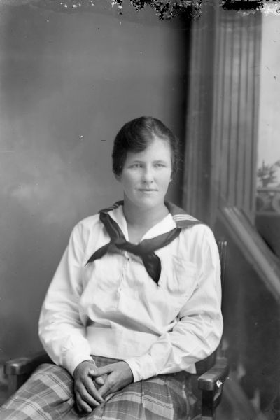 Three-quarter length studio portrait in front of a painted backdrop of an unidentified European American woman posed sitting. She is wearing a plaid skirt, light-colored sailor blouse, and loose necktie.