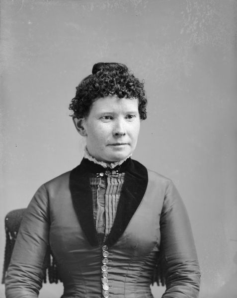 Waist-up studio portrait of an unidentified European American woman posed sitting in a chair with a tasseled back. She is wearing a dark-colored dress with a button-down bodice, pleated front with dark lapels, and a collar pin.