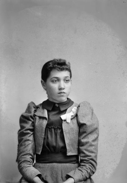 Waist-up studio portrait of an unidentified European American girl posed sitting. She is wearing a light-colored skirt, short jacket, a dark-colored blouse, ribbon and lapel pin, and iron-cross-shaped collar pin.
