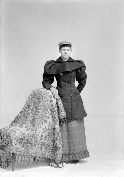 Full-length studio portrait in front of a white backdrop of an unidentified European American woman posed standing. She has her right hand on the back of a draped chair on the left, and is wearing a three-quarter length winter coat with puffy sleeves and wide lapels, a plaid skirt with ruffled trim, and a cap.