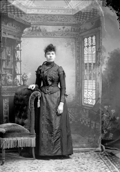 Full-length studio portrait in front of a painted backdrop of an unidentified European American woman posed standing with her right hand on the back of an upholstered chair. She is wearing a dark-colored moiré silk dress with ribbon trim.