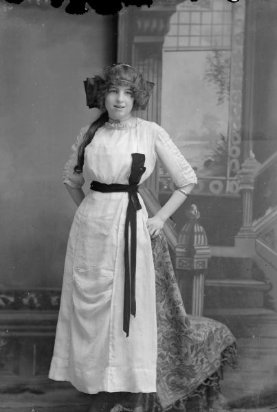 Full-length studio portrait in front of a painted backdrop of an unidentified European American woman posed standing with her hands on the back of a chair behind her. She is wearing a light-colored dress with a ribbon and bow belt/sash, a choker necklace, and a large hair ribbon.