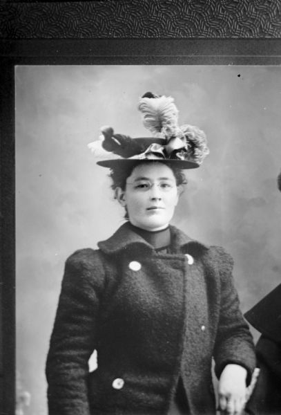 Copy photograph of a waist-up studio portrait of an unidentified European American woman posed standing with her left hand on the back of an object on the right. She is wearing eyeglasses, a dark-colored winter coat and a hat with a ribbon and feathers.