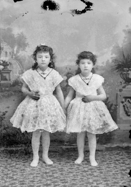 Studio portrait in front of a painted backdrop of two European American girls posed standing and holding hands. They are both wearing matching, light-colored short dresses with lace trim, and necklaces with heart-shaped lockets, collar pins, and tights with no shoes. Identified as probably Ella Odey standing on the left, and Midge Odey standing on the right, both reportedly toe dancers at the Black River Falls Opera House in productions put on by a Chicago firm.
