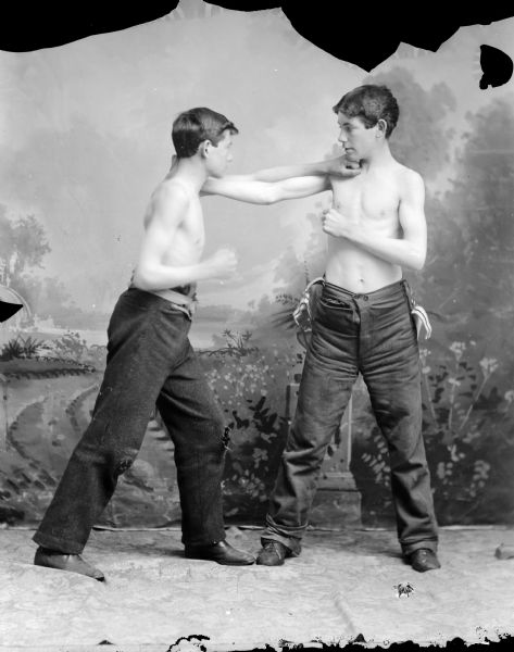 Studio portrait in front of a painted backdrop of two European American boys posed fighting, holding up their right fists as if punching. They are both wearing dark-colored trousers and no shirts.