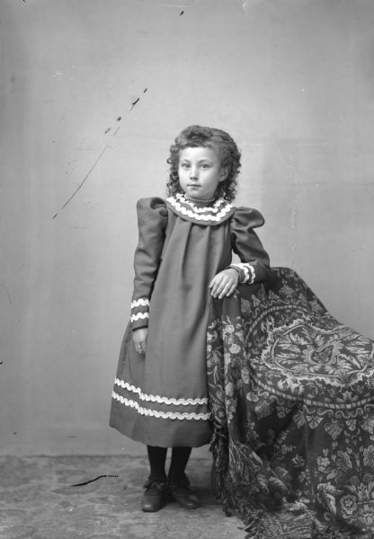 Full-length studio portrait of an unidentified European-American girl posed standing with her right arm resting on the back of a chair draped with a tasseled cloth. She is wearing a three-quarter length light-colored dress with lighter-colored rickrack trim, a choker necklace, and a ring.
