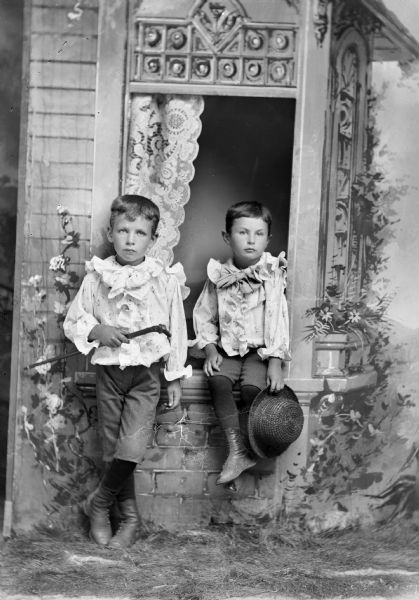 Studio portrait of two unidentified European American boys posed against a prop window sill in front of a painted backdrop. The boy on the left is posed standing and holding a short walking stick in his right hand. He is wearing short, dark-colored knickerbocker pants and a light-colored ruffled blouse with soft neck tie. The smaller boy posed sitting on the right is holding a straw hat in his left hand and wearing short, dark-colored knickerbocker pants and a light-colored ruffled blouse with soft neck tie.