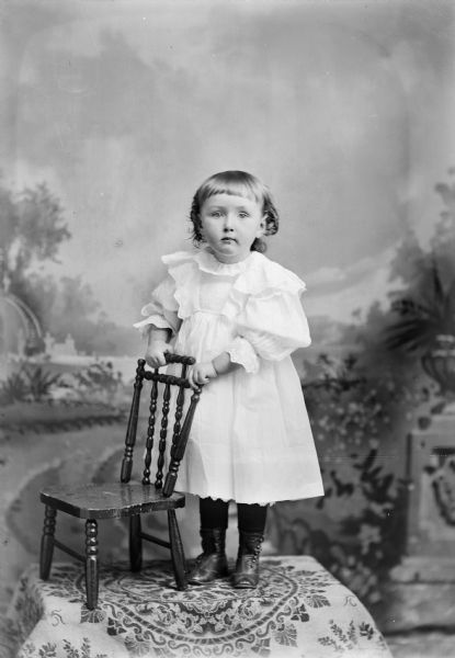 Full-length studio portrait in front of a painted backdrop of a small unidentified European American girl posed standing on top of a table draped with a cloth. She is holding onto the back of a small wooden chair with both hands and is wearing a light-colored dress.