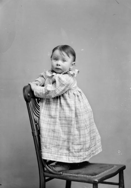 Full-length studio portrait of an unidentified European American girl posed standing sideways on the seat of a chair and holding on to its back with both hands. She is wearing a light-colored plaid dress with rickrack trim.