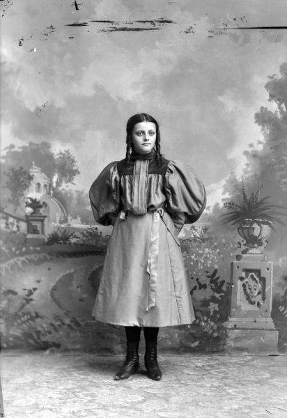 Full-length studio portrait in front of a painted backdrop of an unidentified young European American woman posed standing. She is wearing a light-colored three-quarter length dress with lighter-colored trim.