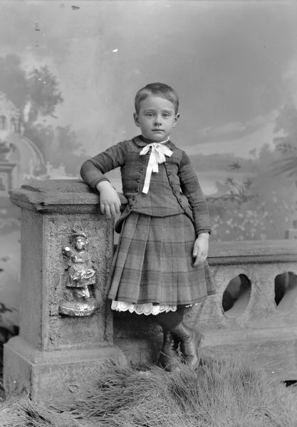 Full-length studio portrait in front of a painted backdrop of an unidentified European American boy posed standing in front of a propr stone wall. He is wearing a plaid dress with a white ribbon around his neck.