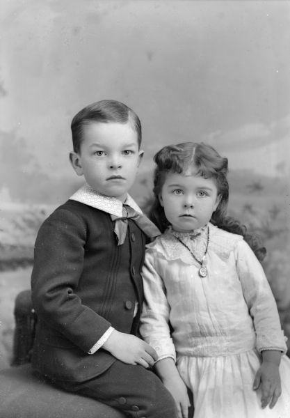 Three-quarter length studio portrait in front of a painted backdrop of two unidentified European American children posing closely together. The boy on the left is posed sitting on the left, and is wearing a dark-colored suit with a light-colored shirt and ribbon bow tie. The girl is posed standing on the right and is wearing a light-colored dress and necklace.