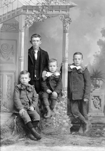 Studio portrait of four unidentified European American boys posed in front of a prop porch setting in front of a painted backdrop. The boy sitting on the left is wearing a dark-colored suit coat, short knickerbockers, and large ribbon bow tie. The second boy and largest child is posed standing with his left hand on the right shoulder of the third boy and is wearing a dark-colored suit coat, vest, and light-colored necktie. The second boy from the right is posed sitting on the porch railing and is wearing a dark-colored suit coat, short knickerbockers, and large light-colored bow tie. The boy on the right is posed standing with his right hand on a wooden post, and is wearing a dark-colored suit coat, short knickerbockers, and large light-colored bow tie.