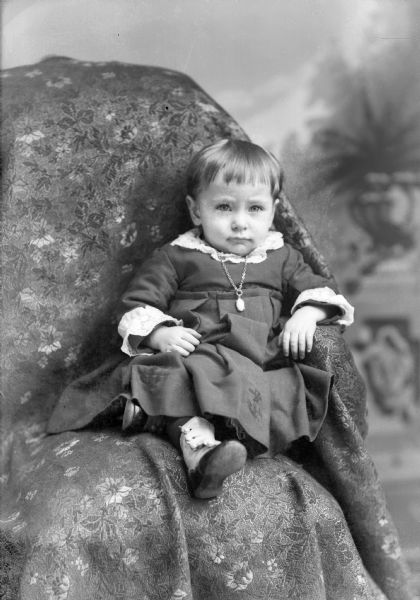 Studio portrait of a small child with short hair posing sitting with their legs up on the seat of a chair draped with fabric. The child is wearing a dark-colored dress with light-colored trim with embroidered anchor design, a chain necklace and locket, and button-up shoes.