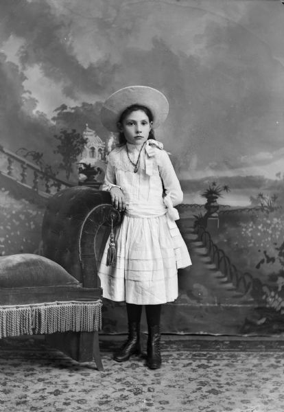 Studio portrait in front of a painted backdrop of an unidentified European American girl posing standing with her right hand on the back of an upholstered chair with a tassel. She is wearing a light-colored  dress with a lace collar with a ribbon, a hat, a necklace and button-up shoes.