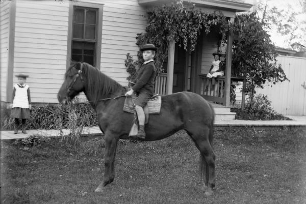 Group outdoor portrait of a boy and two girls posing in front of a wooden house, with foliage growing around the entrance to the door, which has railings and a roof. In the foreground a boy is sitting on a dark-colored horse, wearing dark-colored knickerbockers, suit coat, and hat. On the left is a girl standing on a board sidewalk at the corner of the house. She is blurred from motion and is wearing a light-colored dress and hat. On the right is a girl sitting on the railing near the entrance, wearing a light-colored dress and hat.