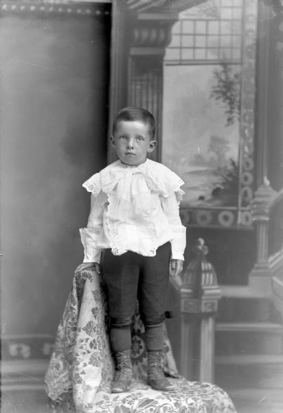 Studio portrait in front of a painted backdrop of an unidentified European American boy posing standing on a chair draped with a cloth. He is wearing a light-colored ruffled shirt with lace trim, and light-colored short knickerbockers.