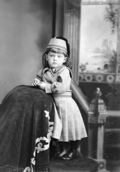 Studio portrait in front of a painted backdrop of an unidentified European American girl posing standing on a platform with her hands resting on the back of a chair covered with a cloth with embroidered trim. She is wearing a light-colored wool or tweed dress, and a matching hat, both with two thin stripes of dark-colored trim.