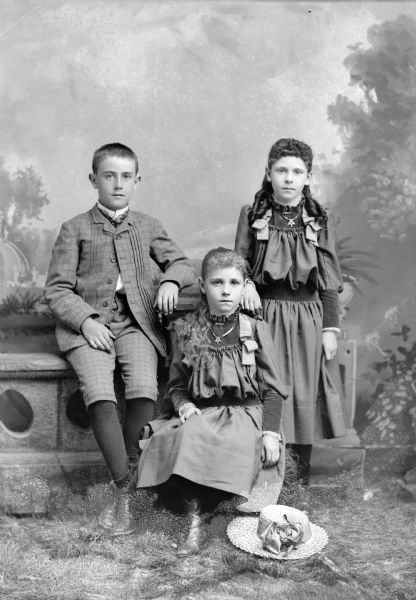 Studio portrait of three European American children. The boy is posing sitting on the left on a stone wall, with his left arm resting on the wall, and is wearing a dark-colored plaid suit coat, knickerbockers, and necktie. The girl sitting lowest on a hay bale and in the center is wearing a dark-colored dress, necklace, and has a straw hat at her feet. The girl posing standing on the right with her right arm on the stone wall is wearing the same type of colored dress and necklace as the girl in the center.