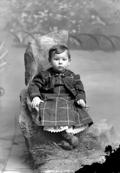 Studio portrait in front of a painted backdrop of an unidentified European American girl with short hair posing sitting on a wooden prop with fake grass at the base. She is wearing a dark-colored plaid dress, light-colored lace slip, and collar pin with a cursive-style letter "L."