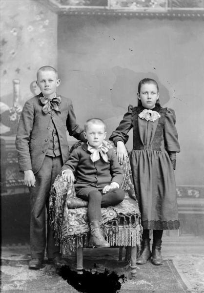 Studio group portrait in front of a painted backdrop of three unidentified European American children. On the left a young man with cropped hair is standing on the left with his left hand on the back of a chair, and wearing a dark-colored suit coat, vest, trousers, and a large plaid ribbon bow tie. A younger boy with cropped hair is sitting on a draped chair in the center. He has his left crossed under his right leg and is wearing dark-colored knickerbockers, sweater, wool tights, and a light-colored neckerchief. On the right is a girl who is standing with her right hand on the chair, and she is wearing a dark-colored dress with lace trim and a light-colored bust ruffle.